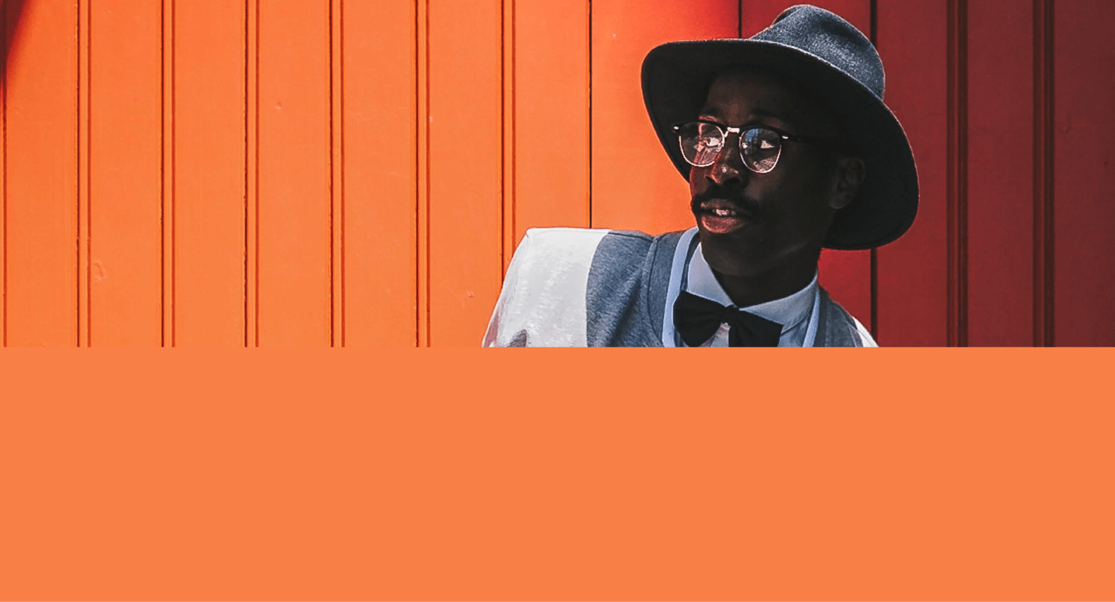 Modelo masculino vestindo chapéu preto, blazer cinza, cammisa social branca, gravata borboleta na cor preta, óculos com lente transparente e armação na cor preta além de um cordão prateado, em um fundo laranja.